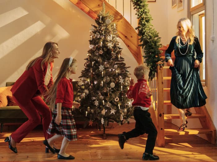 La maman arrive avec les enfants rendre visite à la grand-mère, les enfants courent vers elle dans l’escalier pendant que la grand-mère descend, et à côté de l’escalier se tient le sapin de Noël artificiel en épicéa FairyTrees FT01.