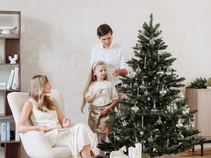 Maman est assise dans un fauteuil clair avec une tasse, tandis que papa et la fille décorent le sapin de Noël artificiel FairyTrees FT14 avec des boules argentées dans le salon.