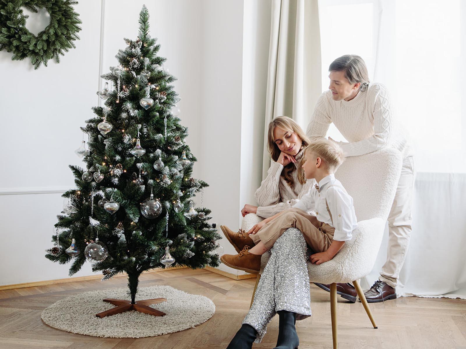 Maman se repose dans un fauteuil avec le fils, papa se penche vers eux, et à côté se tient le sapin de Noël artificiel enneigé FairyTrees FT04 avec des guirlandes lumineuses et des ornements en verre.