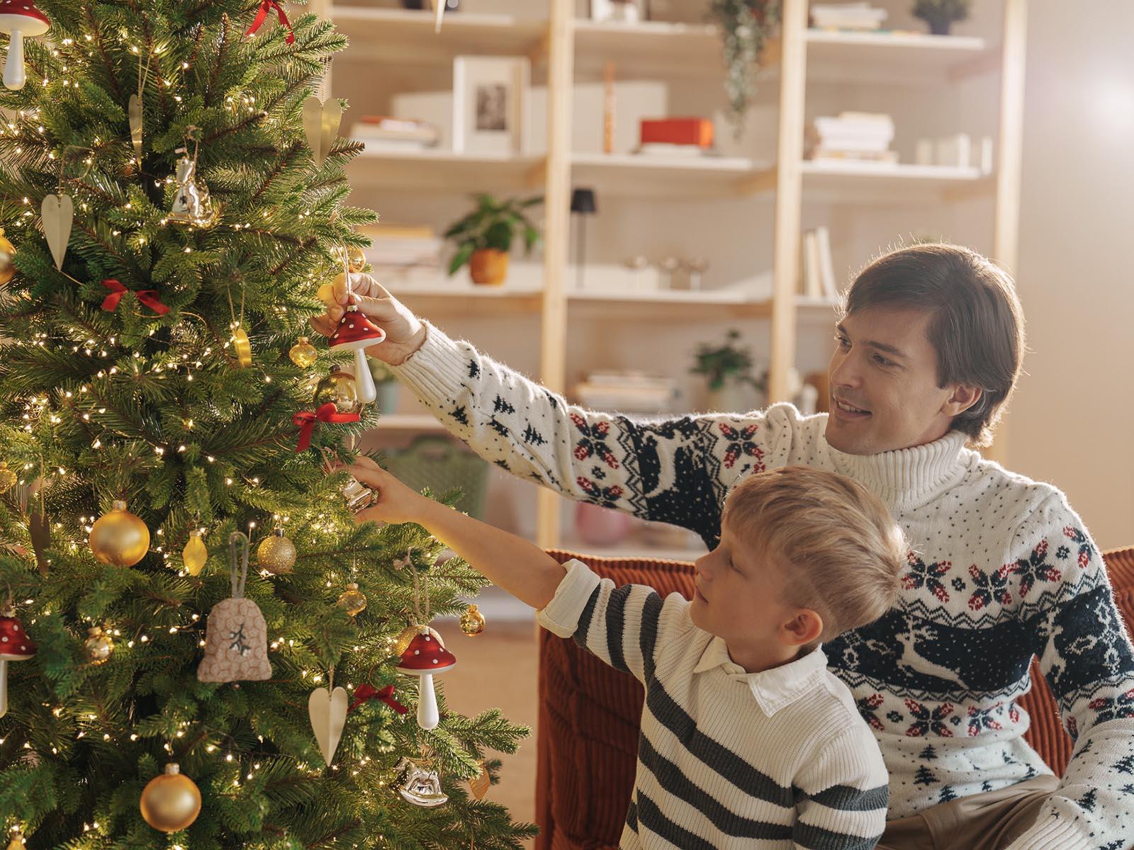 Maman et sa fille décorent le sapin de Noël artificiel FairyTrees FT33 à la lumière chaude du soir.