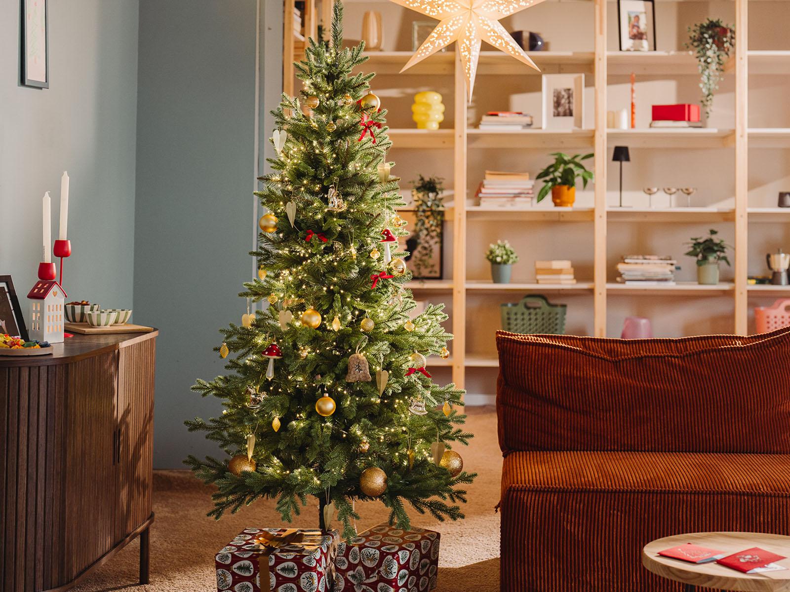 Le sapin de Noël artificiel FairyTrees FT05, un sapin de Nordmann joliment décoré, trône dans le salon d’un petit appartement pendant une soirée d’hiver.