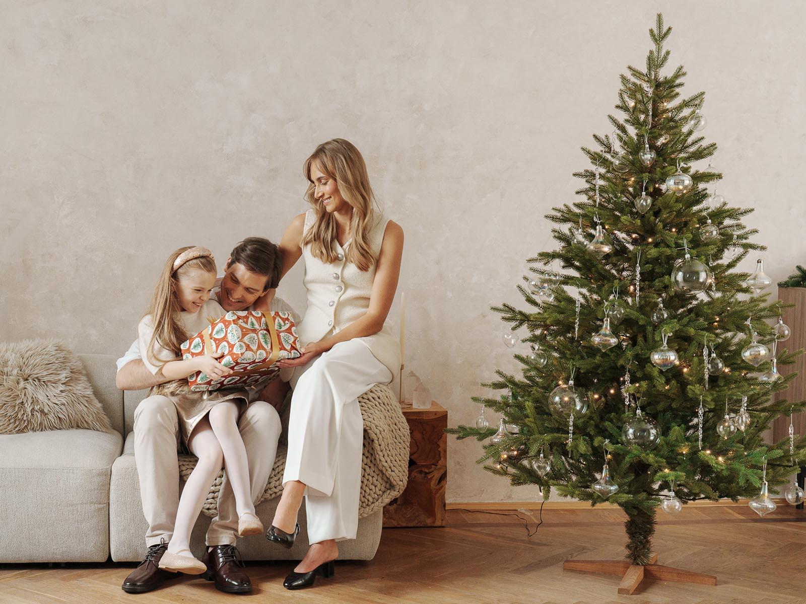 Maman et papa sont assis avec la fille sur le canapé et ouvrent un cadeau, tandis que le sapin de Noël artificiel FairyTrees FT06 brille avec des décorations argentées à côté.