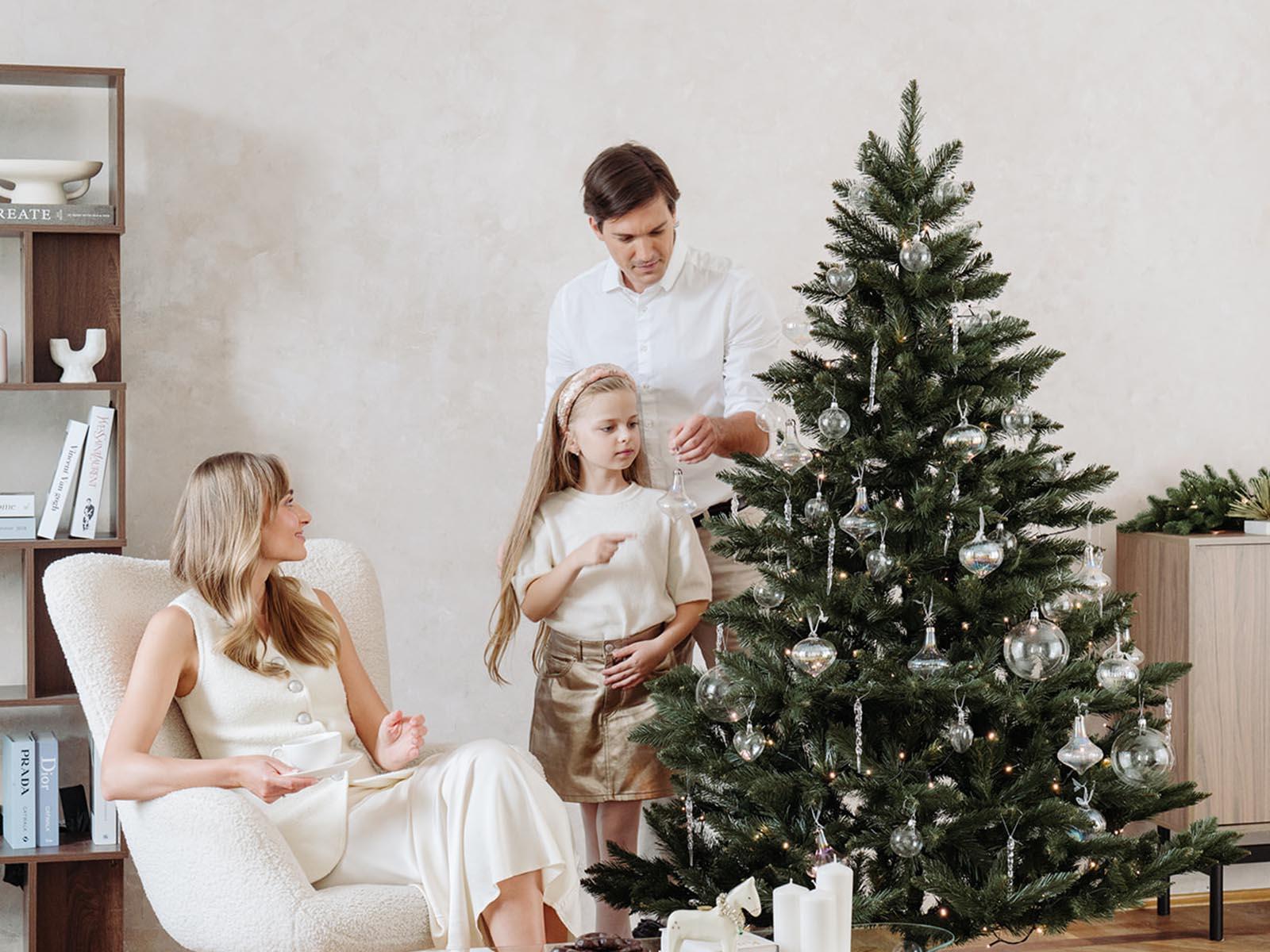 Maman est assise dans un fauteuil clair avec une tasse, tandis que papa et la fille décorent le sapin de Noël artificiel FairyTrees FT14 avec des boules argentées dans le salon.