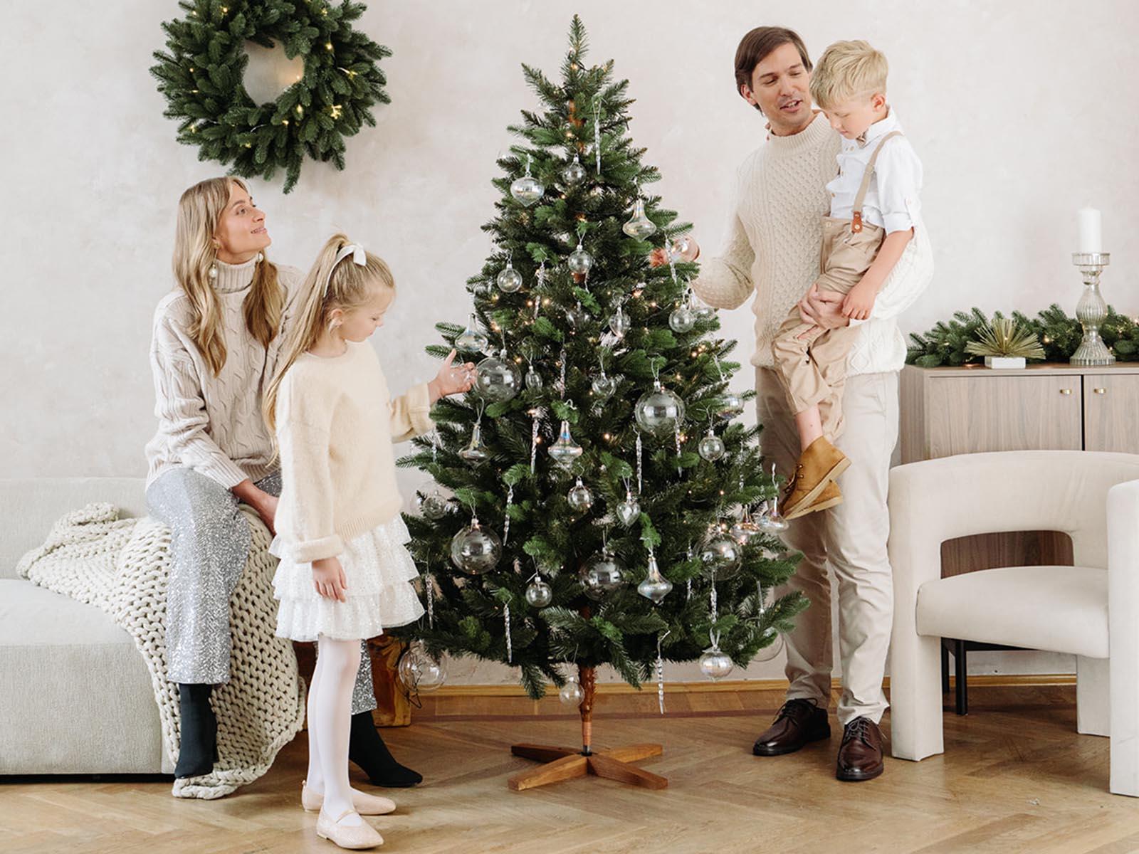 Maman est assise sur le canapé, la fille accroche des décorations et papa porte le fils près du sapin de Noël artificiel FairyTrees FT29 décoré de boules argentées.