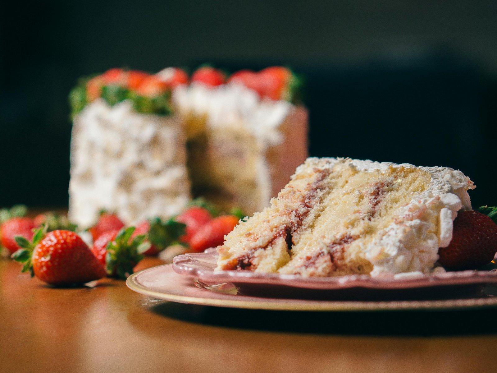 Une part de g&acirc;teau de No&euml;l avec de la cr&egrave;me et des fraises est pos&eacute;e sur une assiette, tandis que le g&acirc;teau entier d&eacute;cor&eacute; est visible en arri&egrave;re-plan.