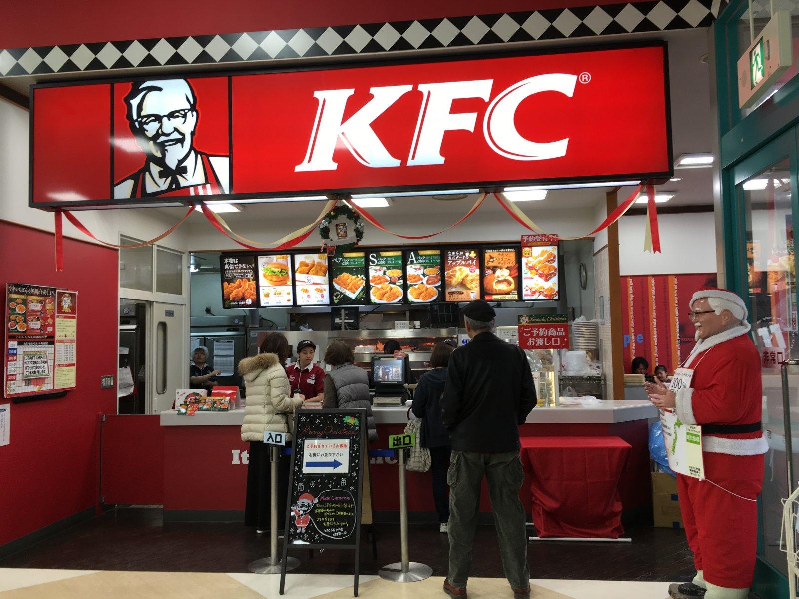Des clients commandent au comptoir d&rsquo;un restaurant KFC d&eacute;cor&eacute; pour No&euml;l, avec une personne d&eacute;guis&eacute;e en P&egrave;re No&euml;l &agrave; c&ocirc;t&eacute;.